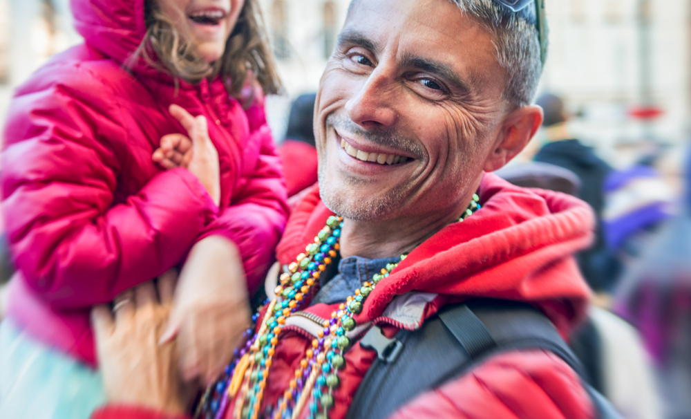 Father in a red hoodie adorned with multiple beaded necklaces hoists his young laughing daughter into the air.