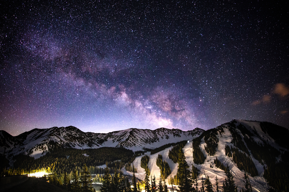 The Milky Way clearly visible above a lit-up Arapahoe Basin mountain out night, its trails illuminated.