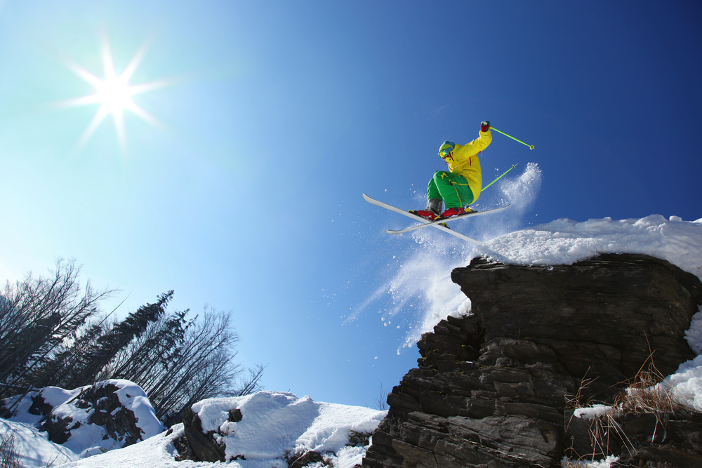 Skier in a yellow parka and green snow pants jumping off a cliff with blue sky behind him.