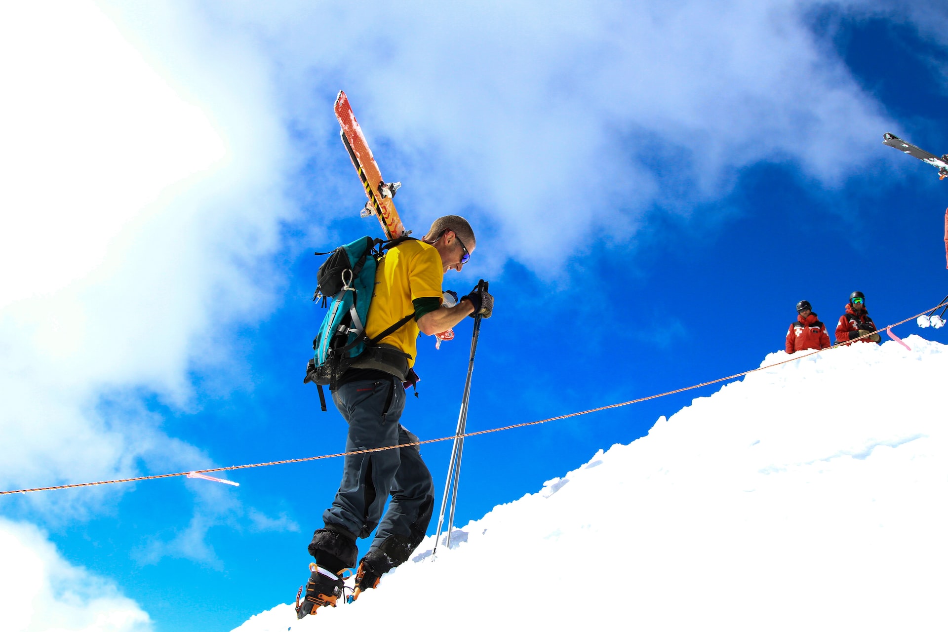 A man walking with skis over his shoulder up a mountain with a blue sky background.