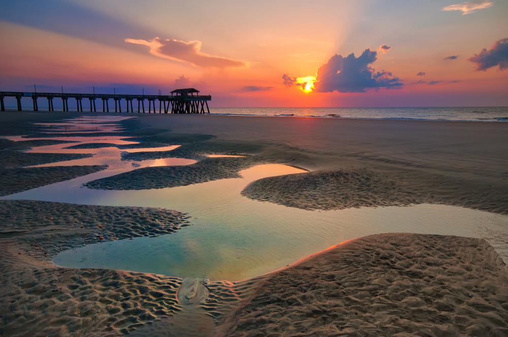 Tybee Island sunrise causing a rainbow affect on standing water at the beach, with a pier and clouds in the distance partially obscuring the sun.