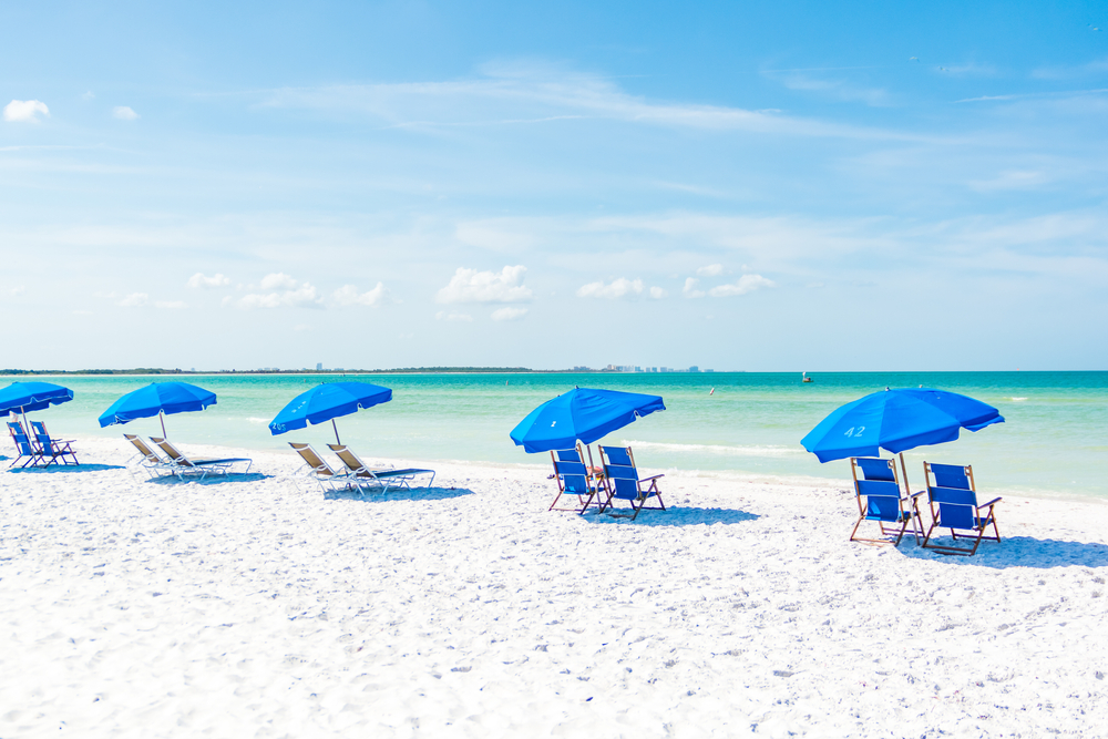 Beachfront Tampa area in Florida; scene showing ocean, white-sand beach, and five blue umbrella and double chaise lounge chair combinations.