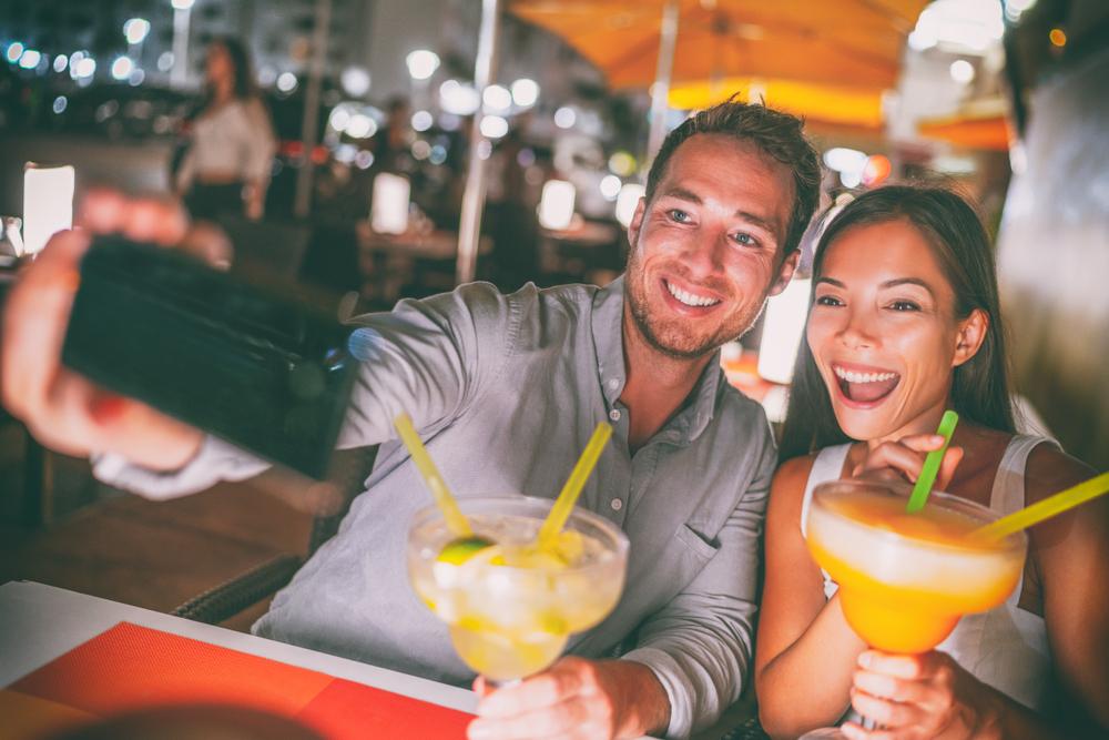 Fun date night out, young couple taking a selfie while drinking giant margarita cocktails while going out at a Miami Ocean Drive restaurant.