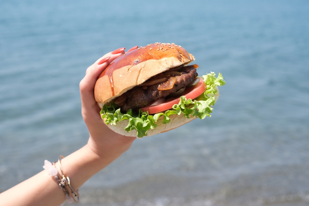 A woman's hand holding up a cheeseburger with the beach in the background.