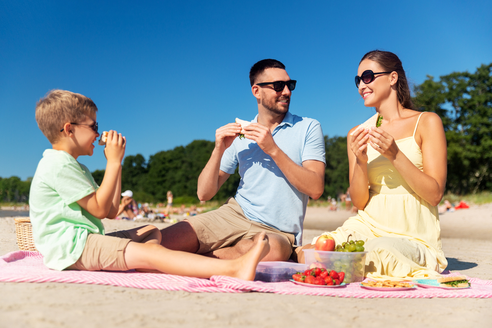 Happy mother, father, and young son having picnic on summer beach and eating sandwiches.