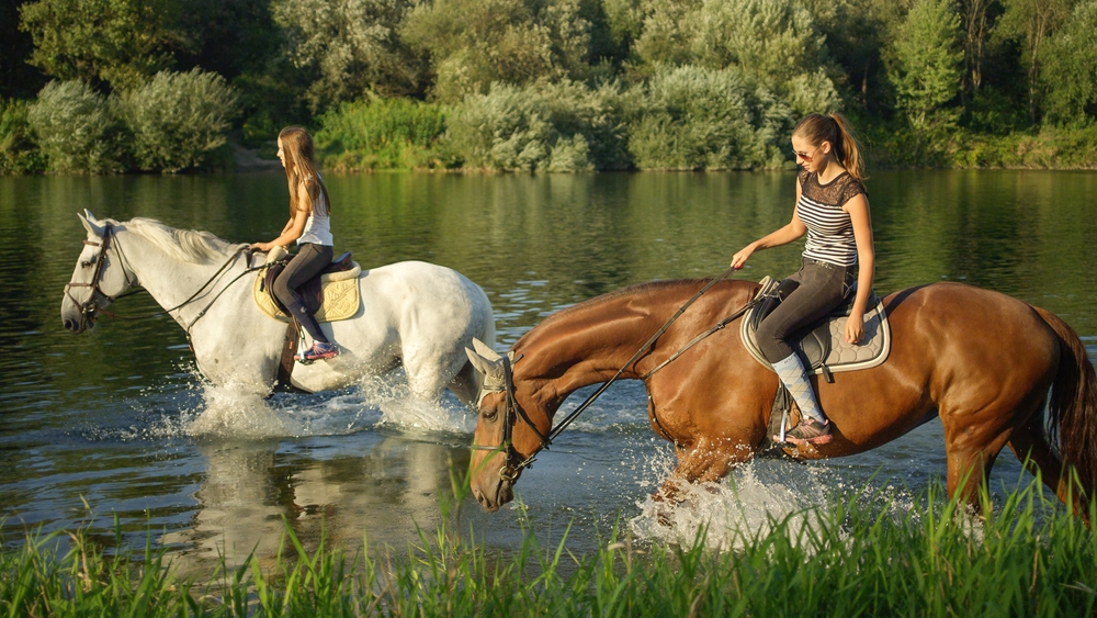 Two girls riding horses along a riverway.