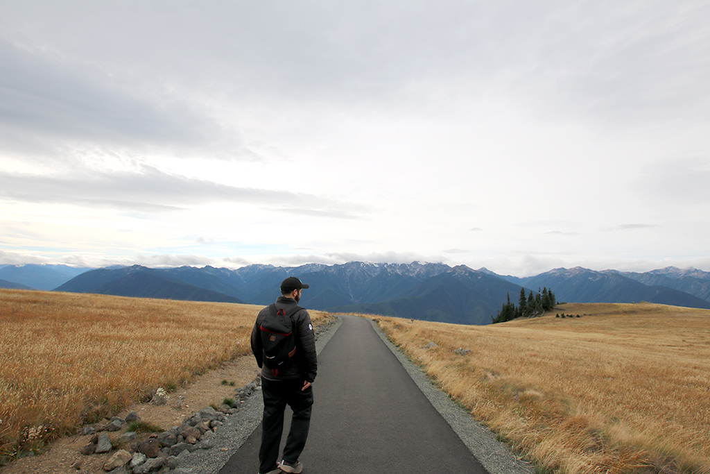 A baseball hat wearing, bearded young man wearing a black jacket and black backpack walks along the road to the Marco Mountains.