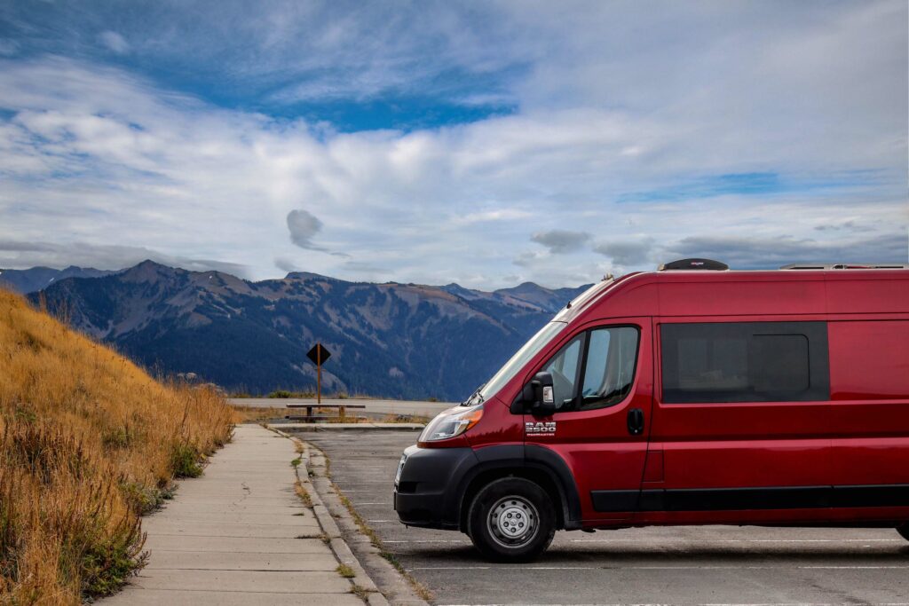 Red camper van parked alone in a parking lot with mountains rising in the distance.