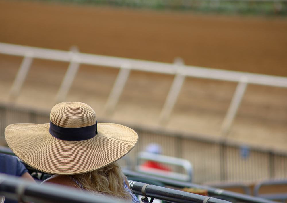 View of a woman with a Derby hat sitting near the dirt track at a horse racetrack with depth of field background.