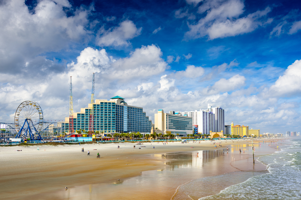 Daytona Beach, Florida, USA beachfront skyline with clouds above, people playing on the beach, and a Ferris wheel visible.