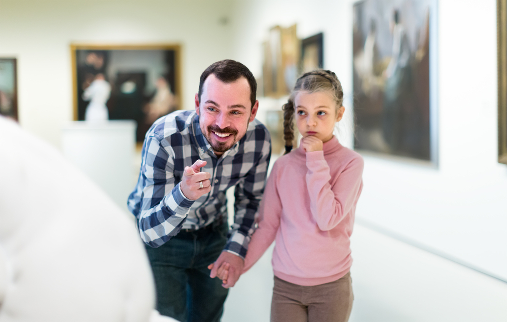 Smiling father and daughter regarding paintings in art exhibit at a museum.