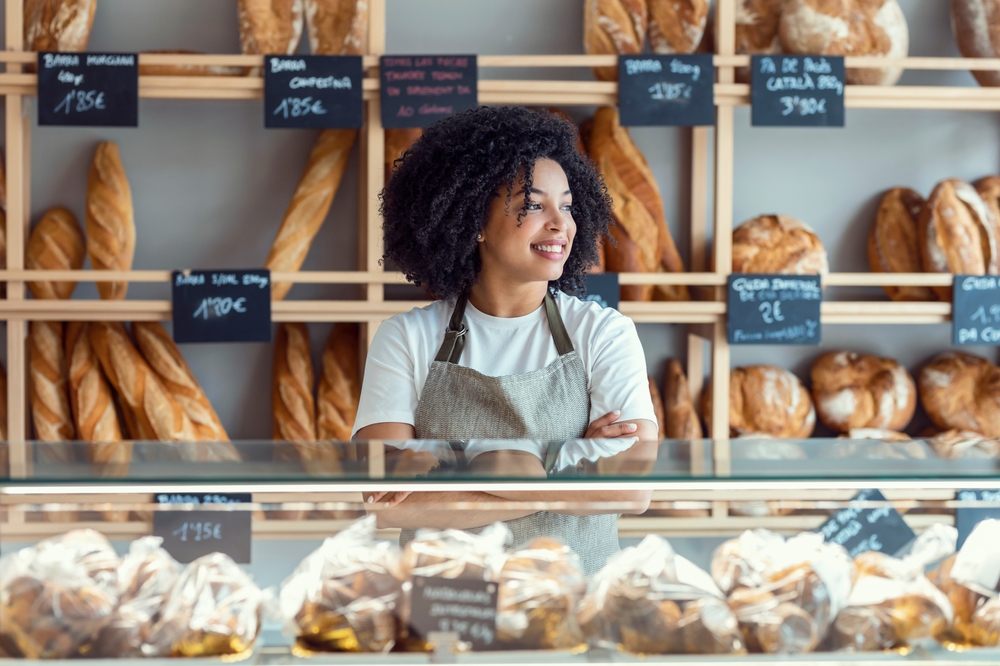 Portrait of beautiful younger owner selling fresh pastry and loaves un bread section and smiling at pastry shop.