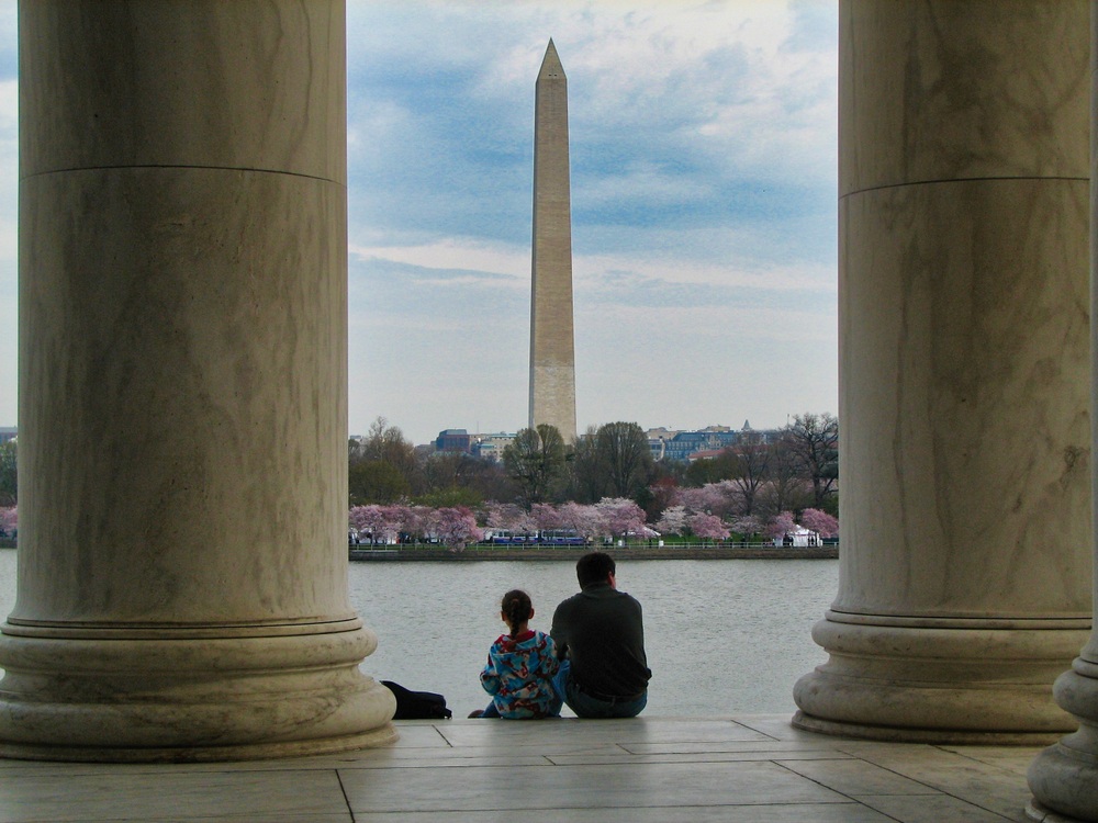 View of the Washington Monument from the Jefferson Memorial with columns and two unidentified people sitting in the foreground.
