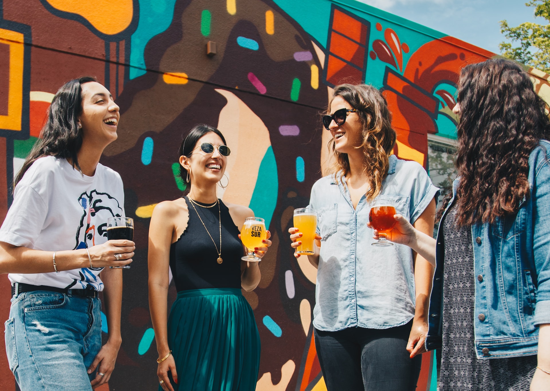 Four women standing around drinking beer in front of a wall painted with graffiti.
