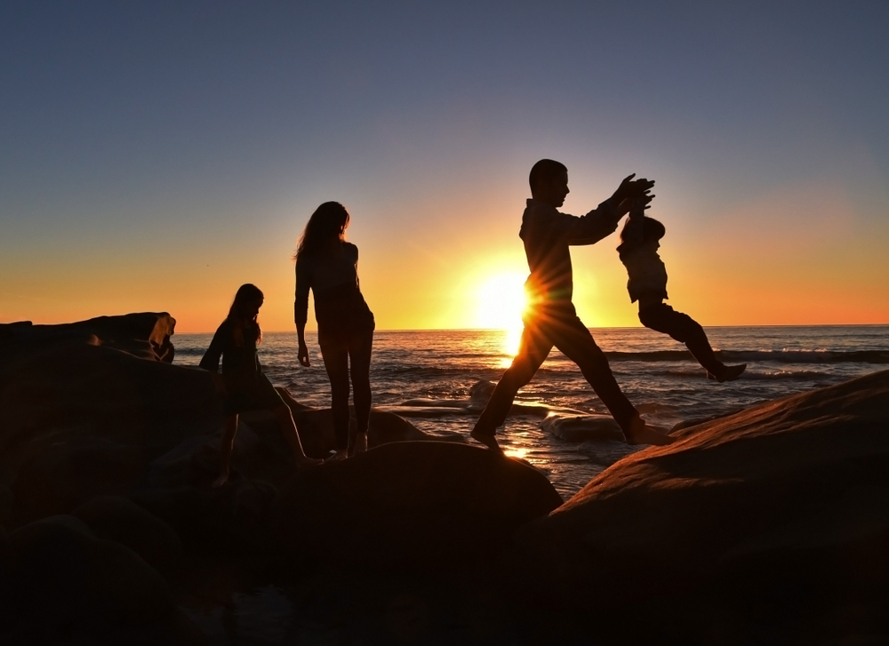 A young family of four enjoying a sunset walk along the cliffs at a San Diego beach. The father is lifting his young son across the boulders.