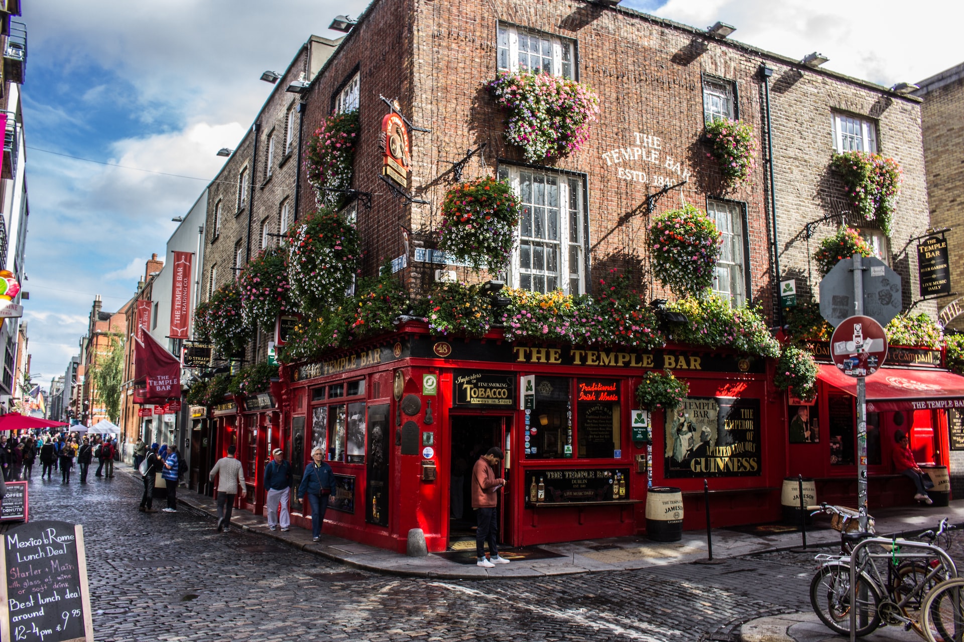 The colorful red Temple Bar on a corner in Dublin with plants hanging from all sides of the building and people standing around outside.