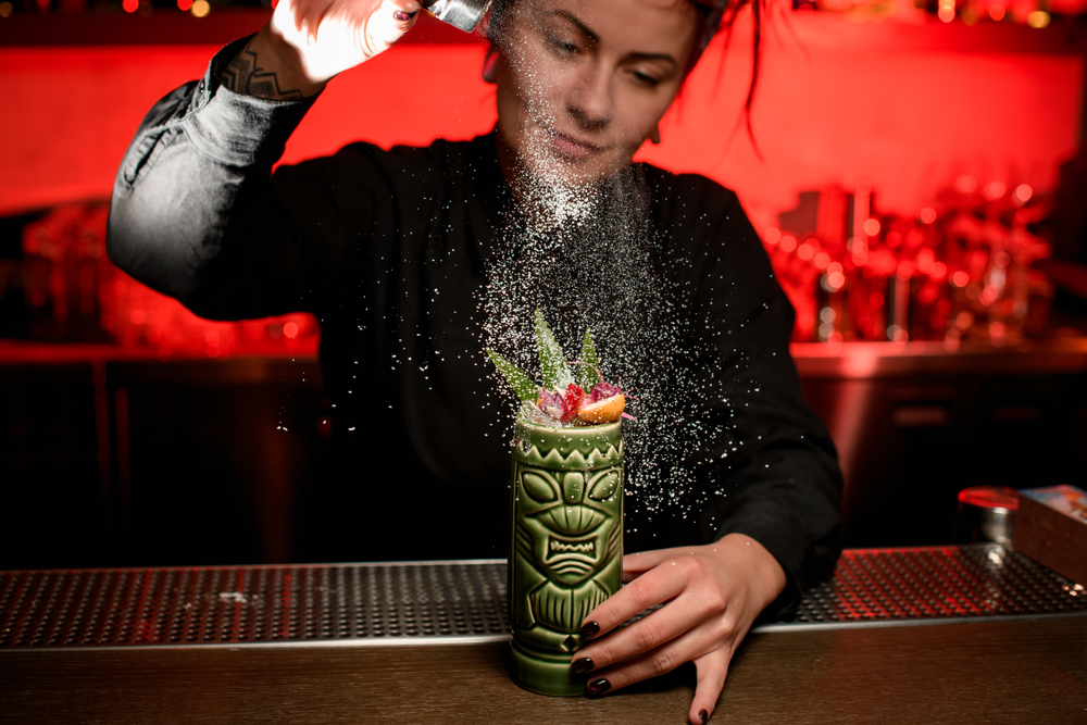 Bartender serving alcoholic cocktail in the Tiki mug sprinkling on it with sugar powder on the steel wooden bar counter.