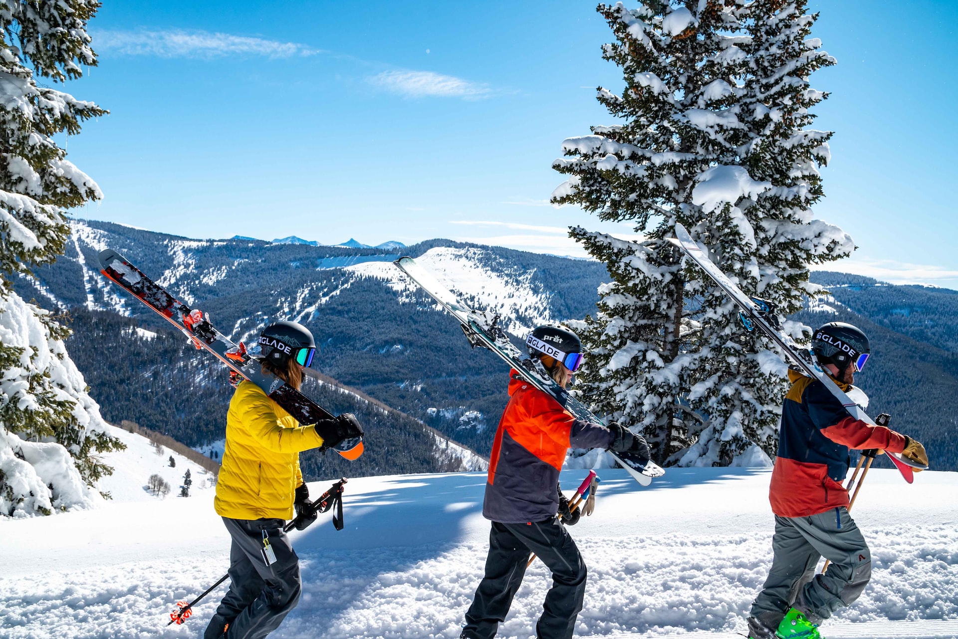 Three skiers in colorful outfits walking with skis over their shoulders across a snowy ridge.