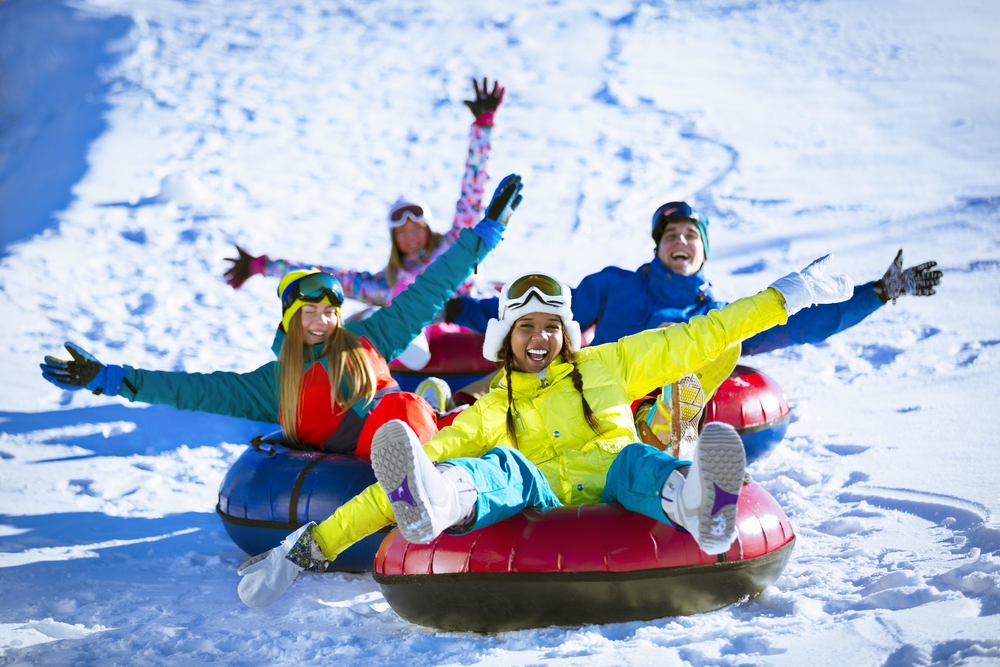 Four happy younger people waving their hands as they go snow tubing down a mountain.