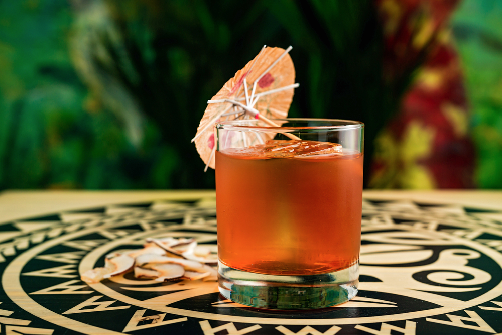 A bright orange tiki-style cocktail in a rocks glass with an ice cube sitting on the table with a miniature umbrella.