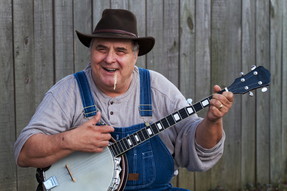A banjo player smiling with a piece of wheat in his mouth wearing a cowboy hat and blue overalls.