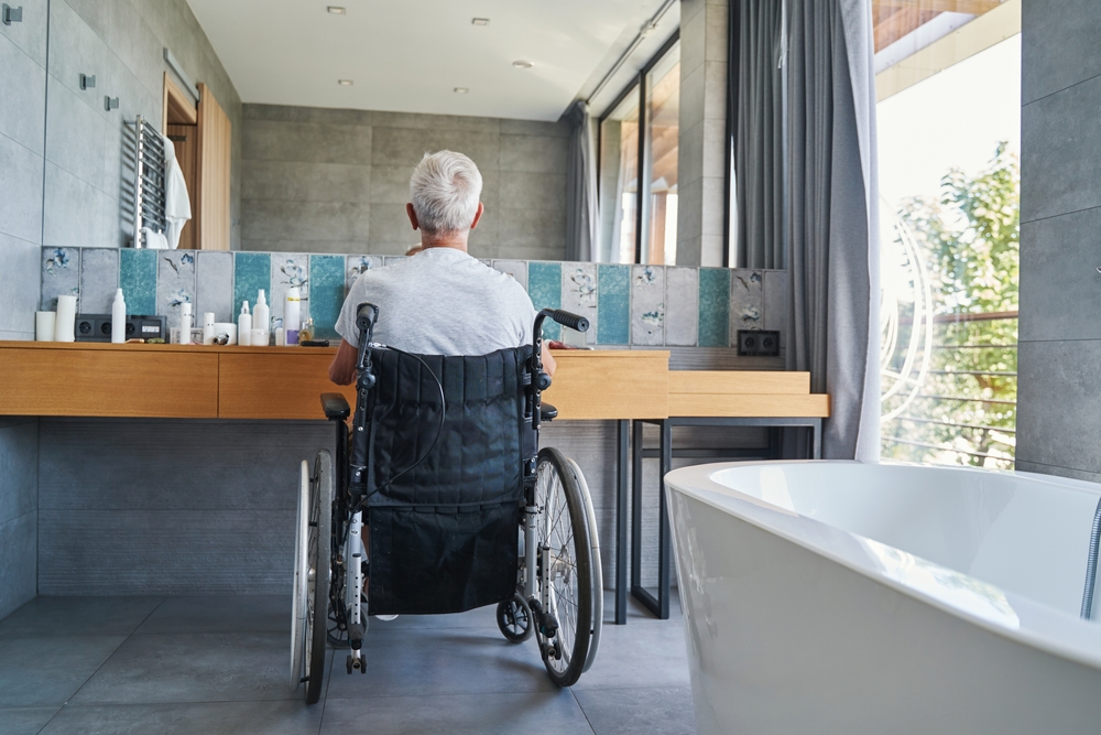Back view of old man in wheelchair before bathroom mirror.