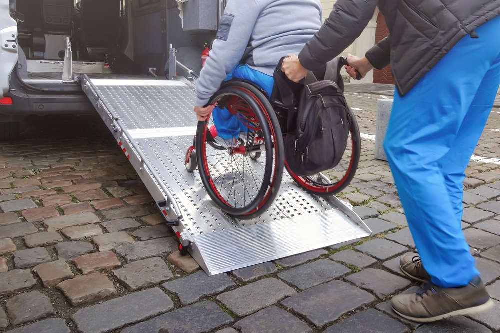 Person in wheelchair with disability using accessible car ramp for transport with help of an assistant driver.