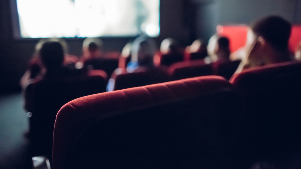 Small movie theater background; audience sitting in the dark watching a movie.