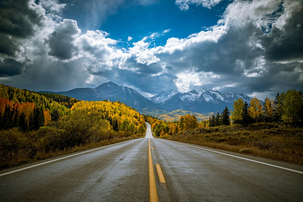 Colorado scenic byway drive, San Juan Skyway after a rainstorm with dramatic clouds and sun illuminating yellow Aspen trees during peak fall color, snow covered mountains in the distance.