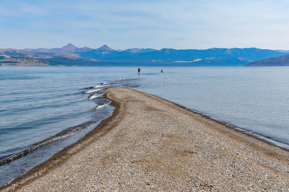 A sandy stretch of Frank Island on Yellowstone Lake with mountains rising in the distance and two people walking into the water.