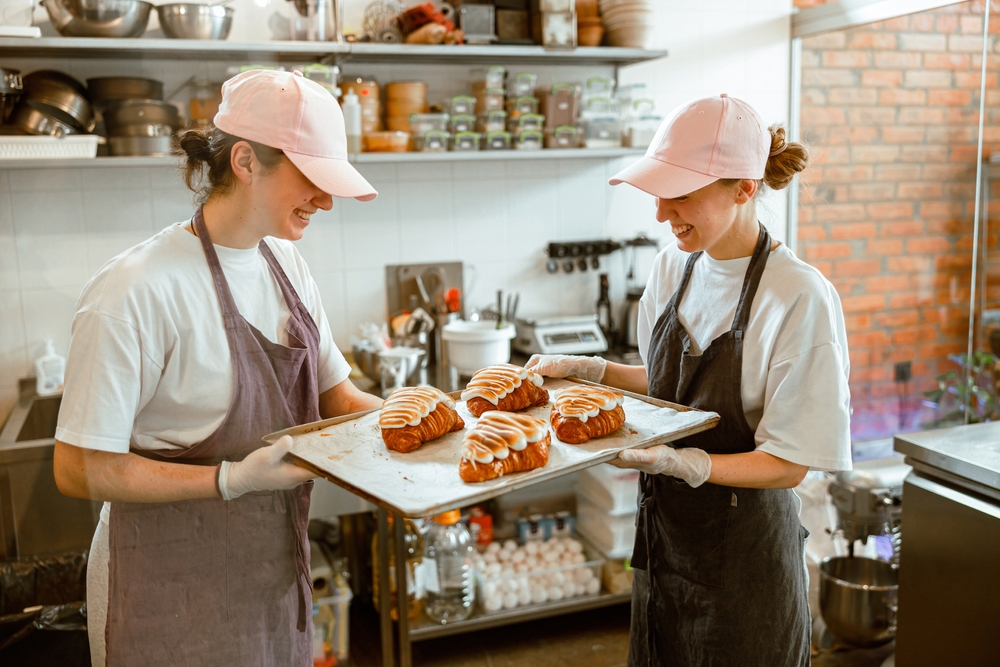 Two smiling women confectioners wearing baseball caps hold a large tray with decorated croissants in a bakery shop.