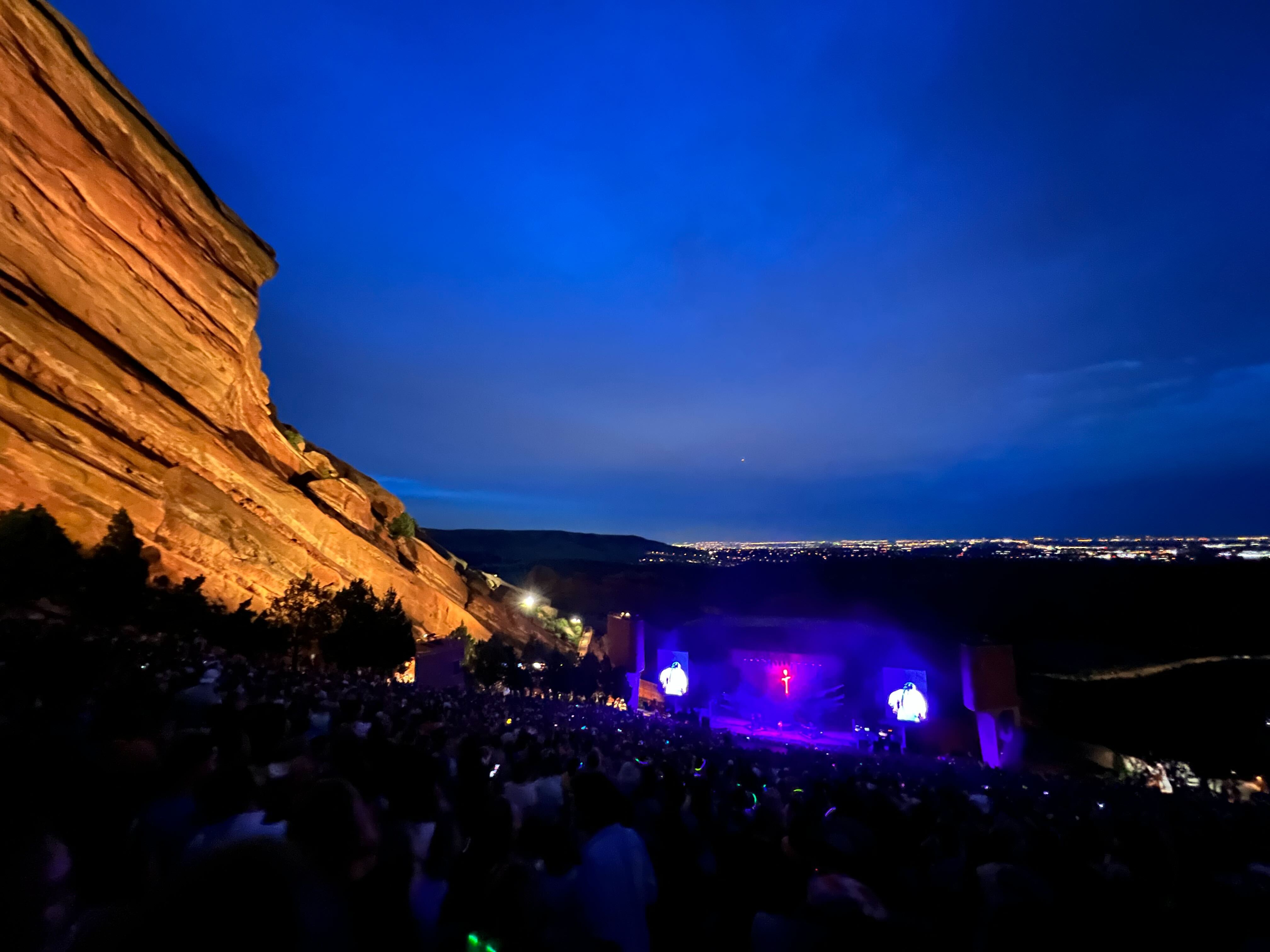 Red Rocks Amphitheater lit up at night, the shot taken from high up of a band performing on stage and the city of Denver lit up in the distance.