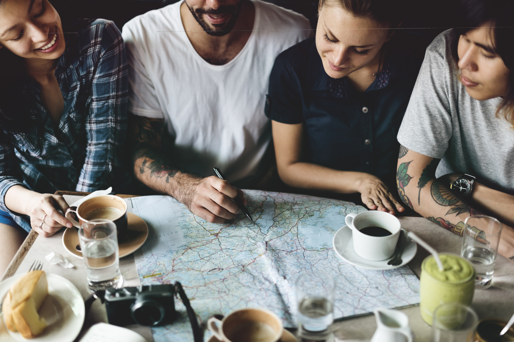 A group of four friends enjoy some coffee while planning out their next adventure with a map spread out between them on the table.