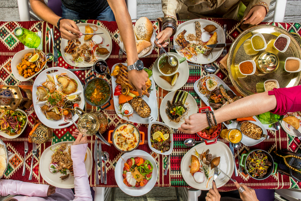 Bird's eye view of a table with many small plates arrayed around with multiple disembodied arms and hands reaching for food.