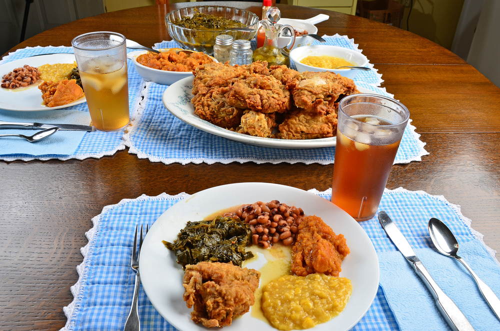 Southern soul food on beautiful old oak dinner table. Wide angle look at plate of fried chicken, creamed corn, collard greens; candied yams and black eyed peas with serving dishes in background.