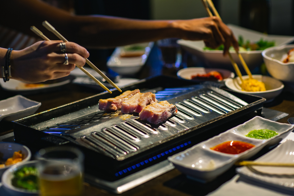 Hands grilling meat via a burner and chopsticks at a Korean BBQ restaurant in Korea Town, Los Angeles.