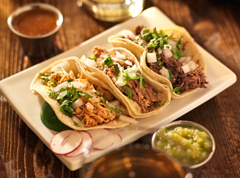 Authentic Mexican barbacoa, carnitas, and chicken tacos served on a rectangular white plate with sides of guacamole and salsa on a wooden table.