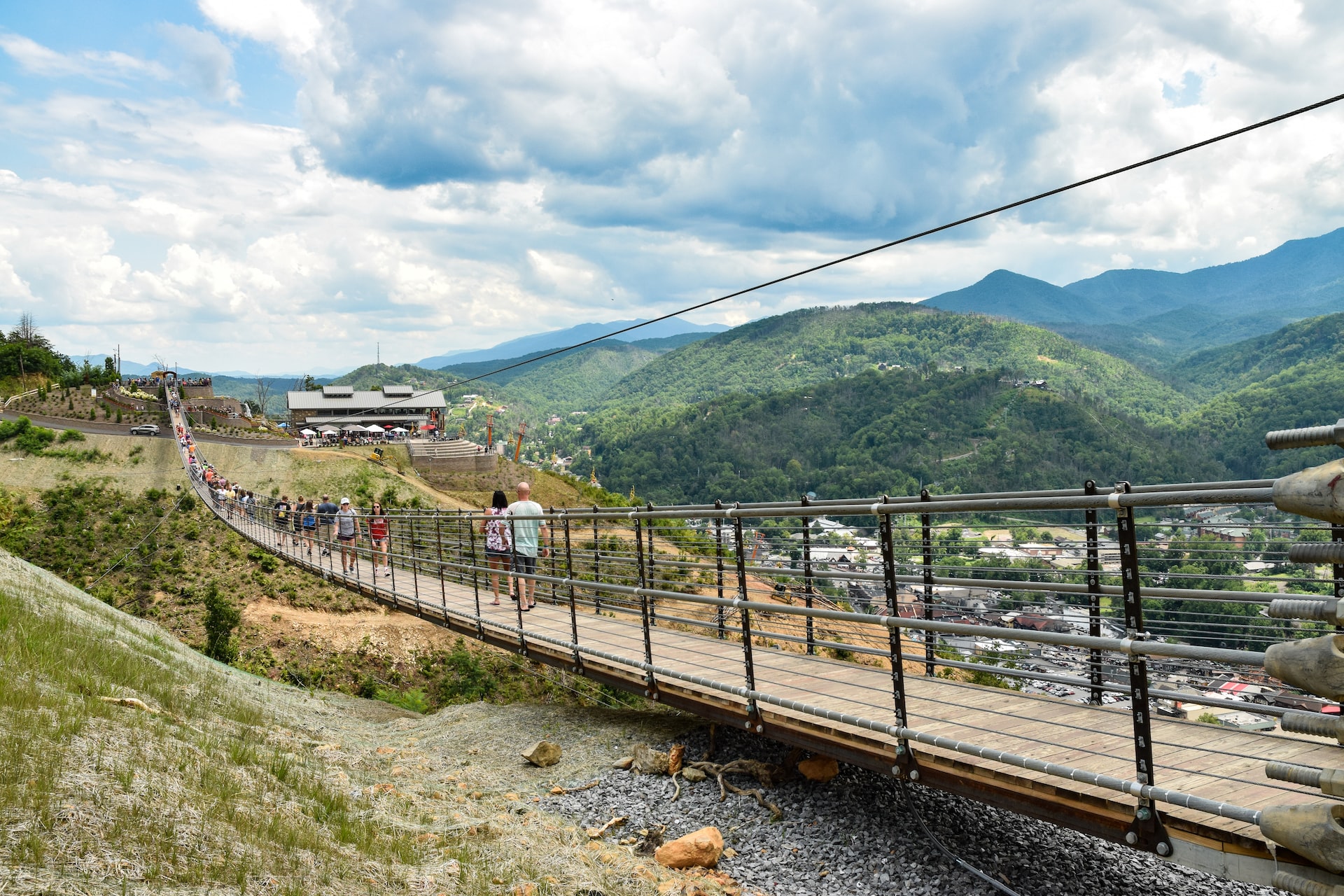 People walking across a narrow hanging bridge with green mountians off in the distance.