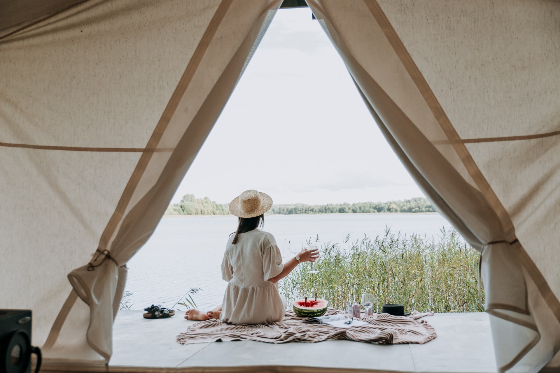 Woman wearing white dress and straw hat visible through the opening in a glamping tent beside a lake.