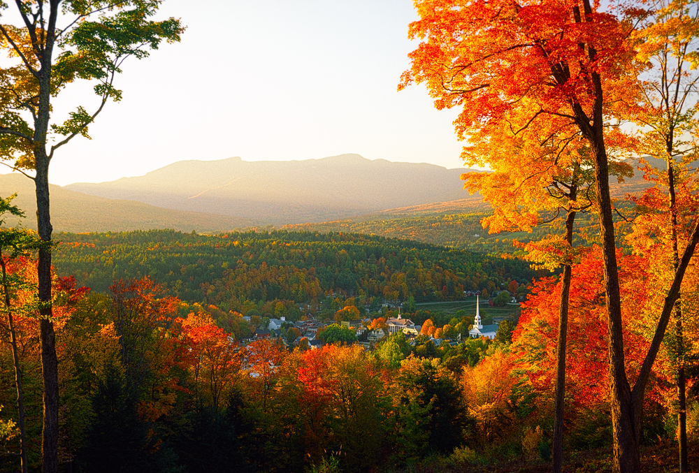 A view framed by foliage covered trees overlooking Stowe Community Church sitting amongst the tranquil village of Stowe on a spectacular autumn sunset with Mt. Mansfield in the background.
