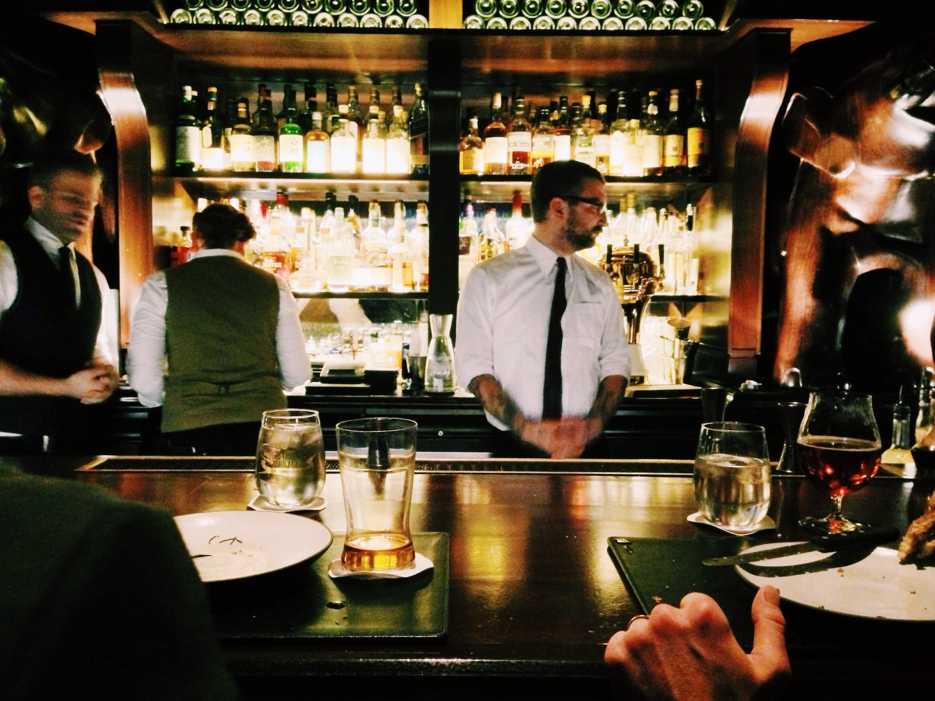 Bartender wearing a black tie and white button down and patrons sitting and standing at the NoMad hotel. 