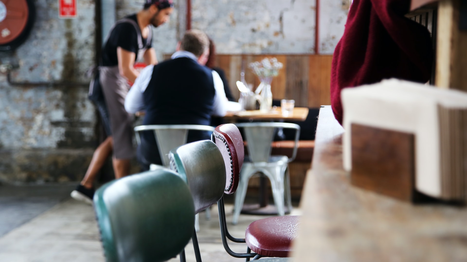 A waiter serving a couple sitting down at the only occupied table, in the foreground are empty barstools.