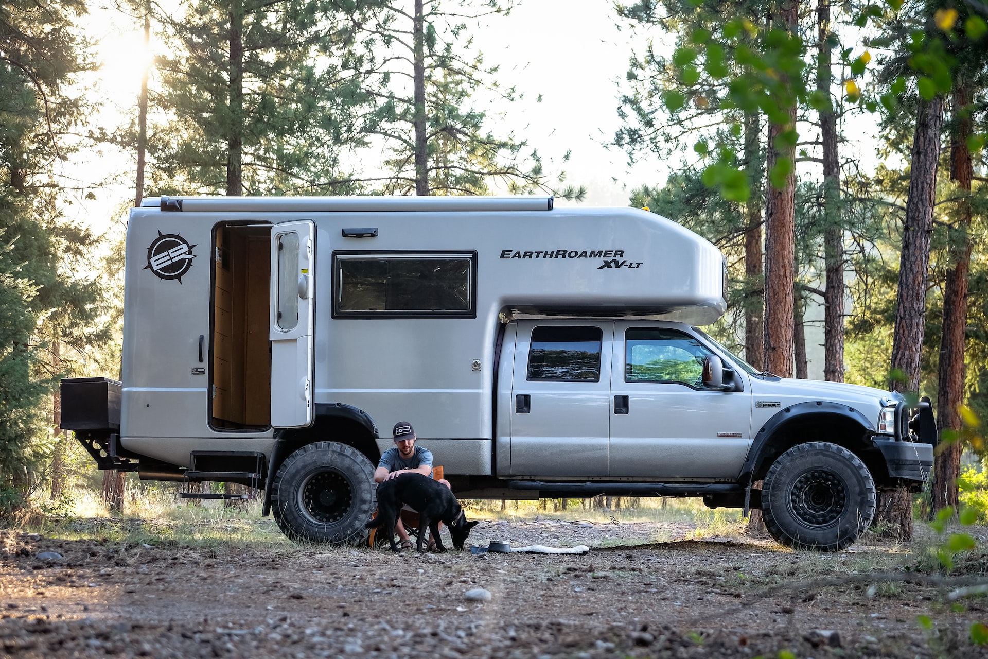 A diesel looking camper in the middle of the woods with a man sitting down near the ground next to his black dog.
