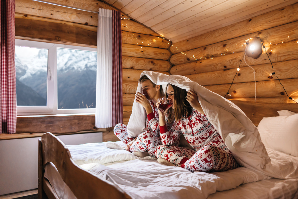 Dreamy Christmas vacation in log house with winter mountains landscape in window. Two teen friends wearing same pajamas drinking tea while relaxing under blanket in bed.