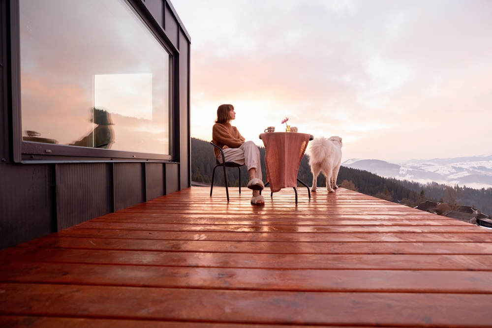 Woman with her dog resting on terrace of tiny house in the mountains, enjoying beautiful landscape during sunrise.