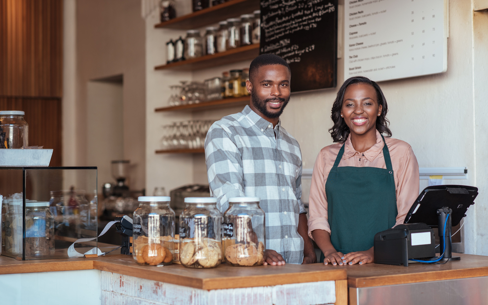 Portrait of two smiling young African-American entrepreneurs standing welcomingly together behind the counter of their trendy cafe.