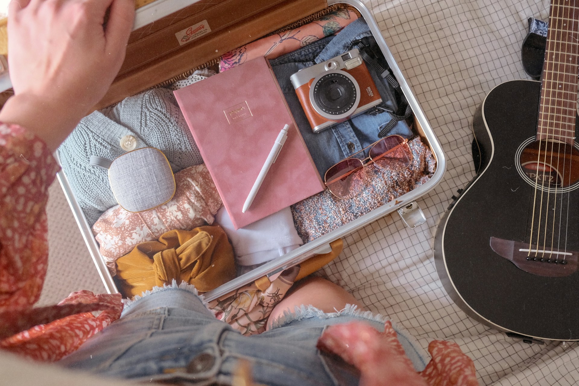 A suitcase being packed from above. A pink notebook and a classic style camera are visible.