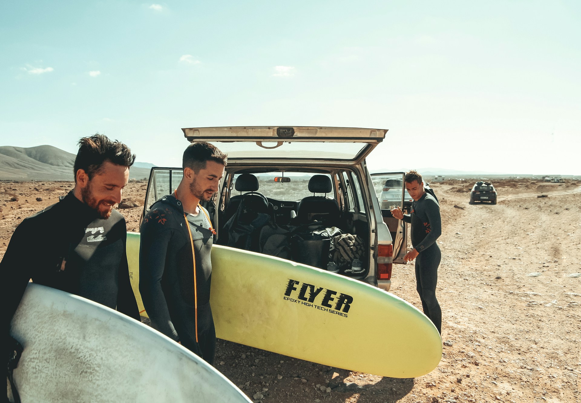 A group of surfers walk by a car they are unpacking with their surfboards in arm