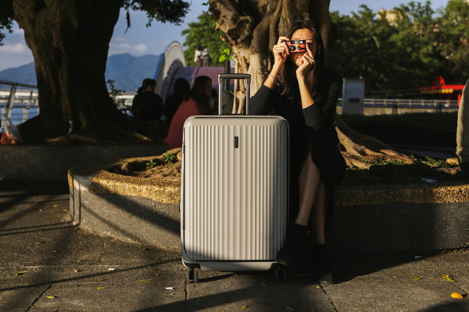 A woman sitting on a park bench, taking a photo through a disposable camera, with a grey suitcase in front of her